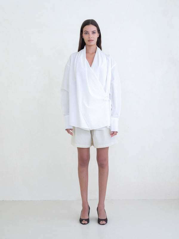 Woman wearing a white wrap shirt and cream shorts on a white background.  